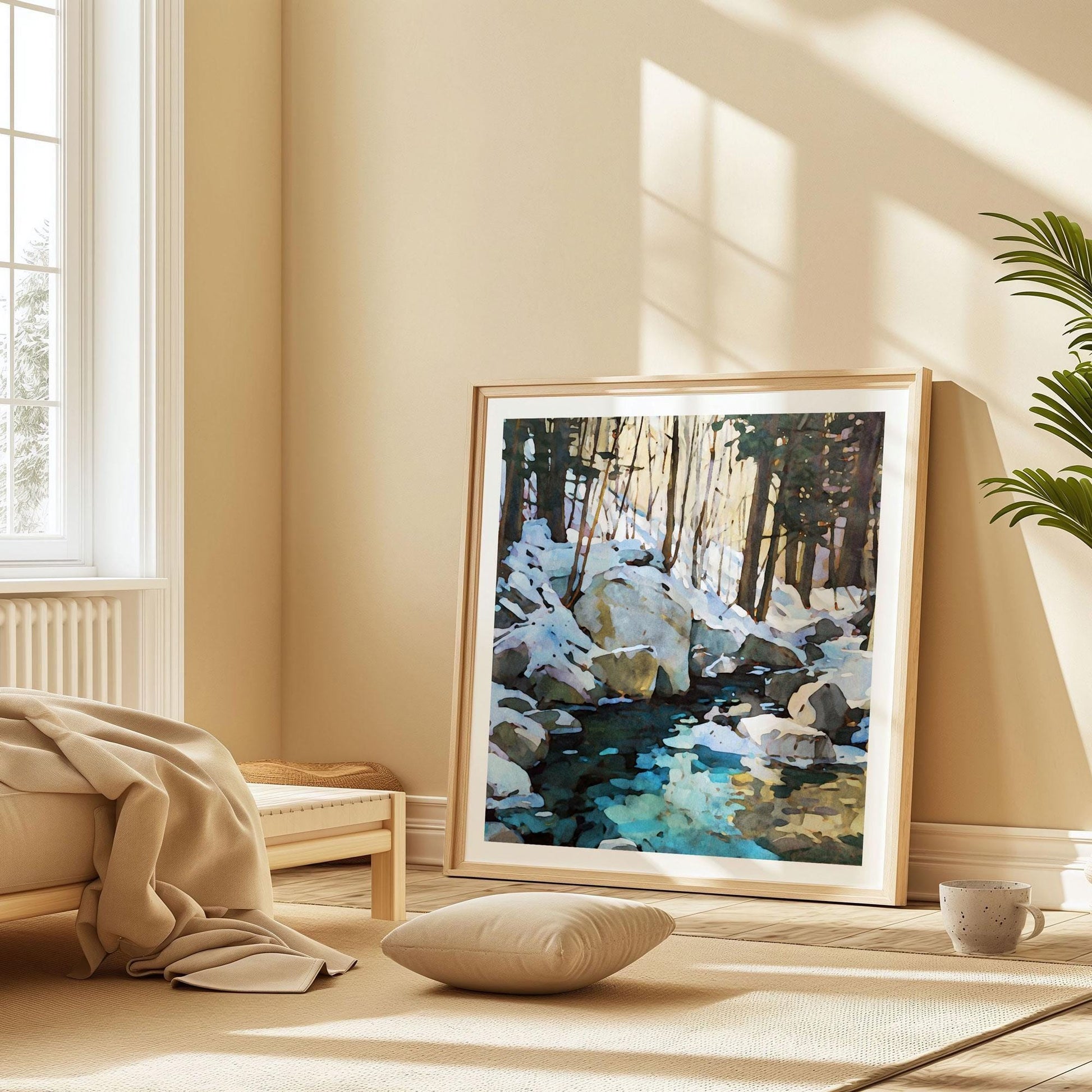 Framed winter forest painting leaning against a beige wall in a sunlit bedroom. The artwork shows snow-covered rocks, tall trees, and a reflective blue-green stream. A low wooden bed with a beige blanket, a floor cushion, and a speckled ceramic mug create a cozy, minimalist setting with warm natural light streaming through the window.
