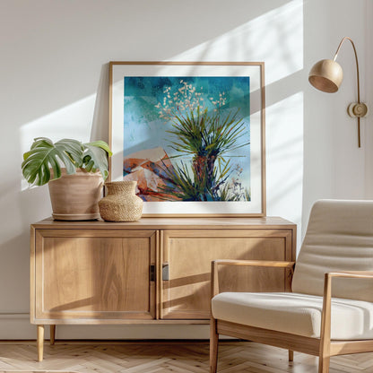 Framed desert yucca painting with spiky green leaves and white blossoms against a turquoise sky and angular orange-blue rocks, displayed on a wooden sideboard. The room features a potted monstera plant, woven basket, neutral armchair, and wall-mounted lamp, with sunlight casting soft shadows across the minimalist, natural-toned interior.