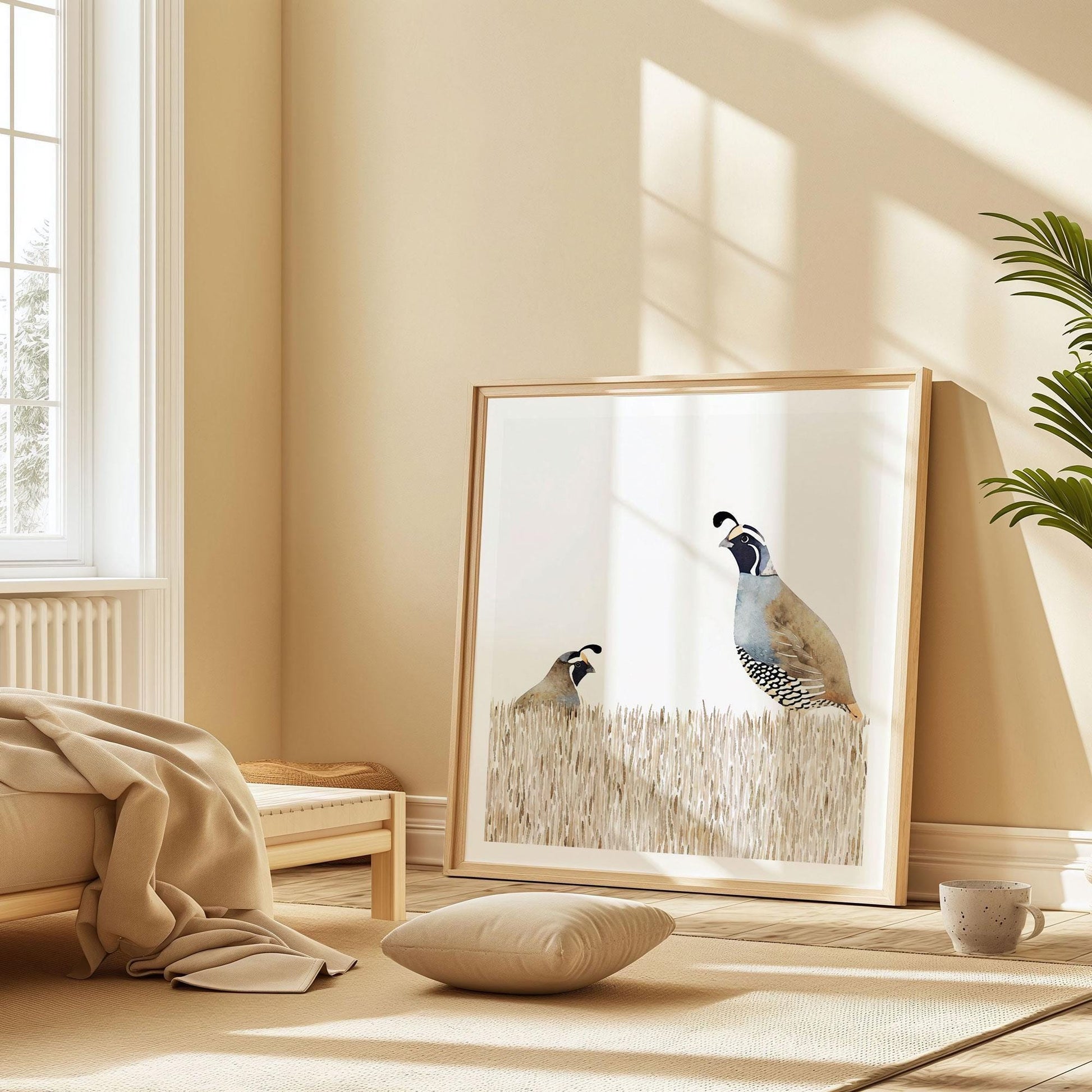 Square framed watercolor print of two California quail in beige grass rests against a warm neutral wall in a sunlit bedroom, styled with a low wooden bed, soft blanket, floor cushion, and a coffee cup on the floor for a cozy minimalist look.