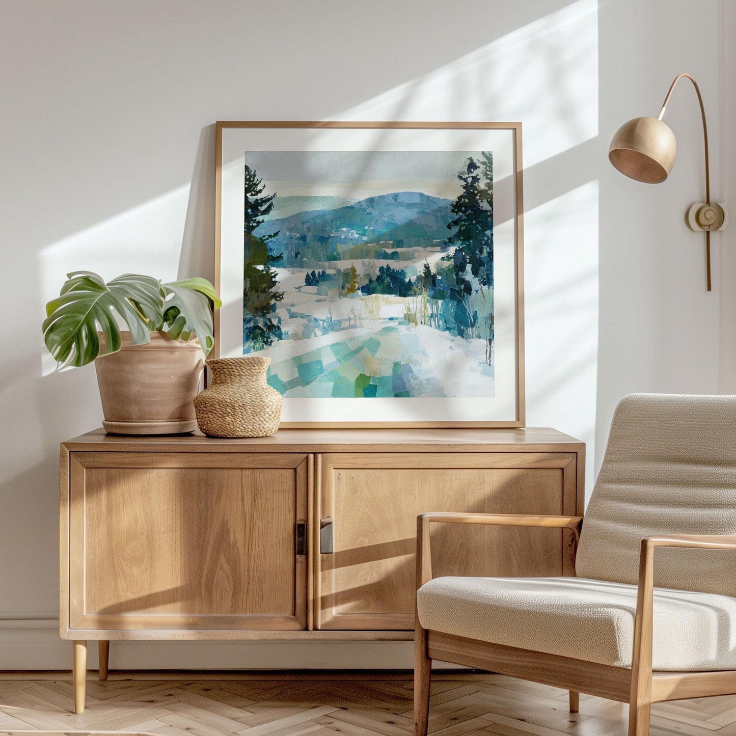 Framed winter mountain landscape painting with snowy hills, evergreen trees, and blue-green tones, displayed on a wooden sideboard with a potted plant and basket, in a bright modern room with natural light and a neutral armchair.