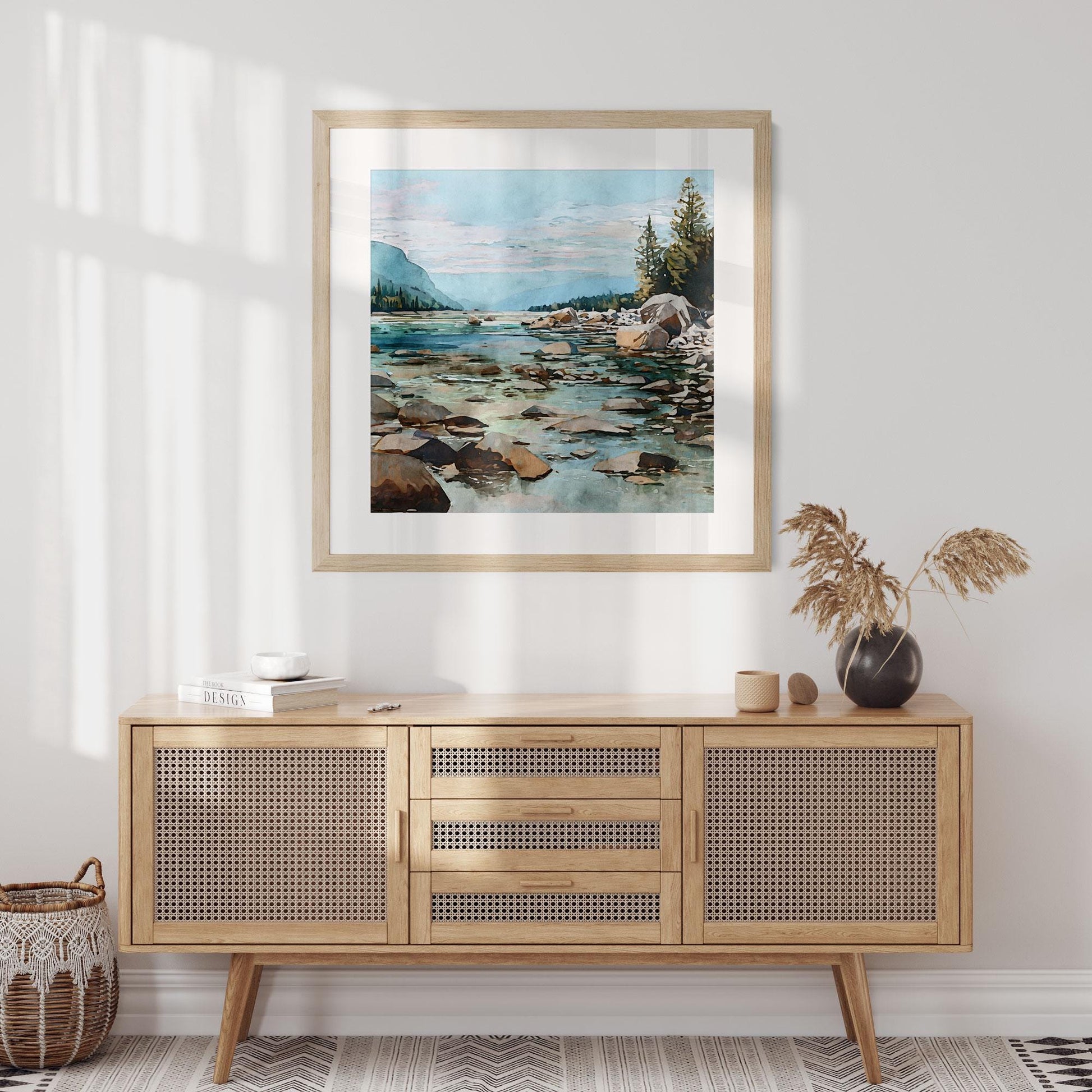 Framed watercolor painting of a rocky mountain river hangs above a mid-century style cane-front sideboard in a bright, minimalist room. The artwork depicts turquoise water flowing through stones toward distant blue mountains and pine trees under a pastel sky. Decor includes a small black vase with dried grass, neutral ceramics, and a woven basket, all softly lit by natural sunlight.