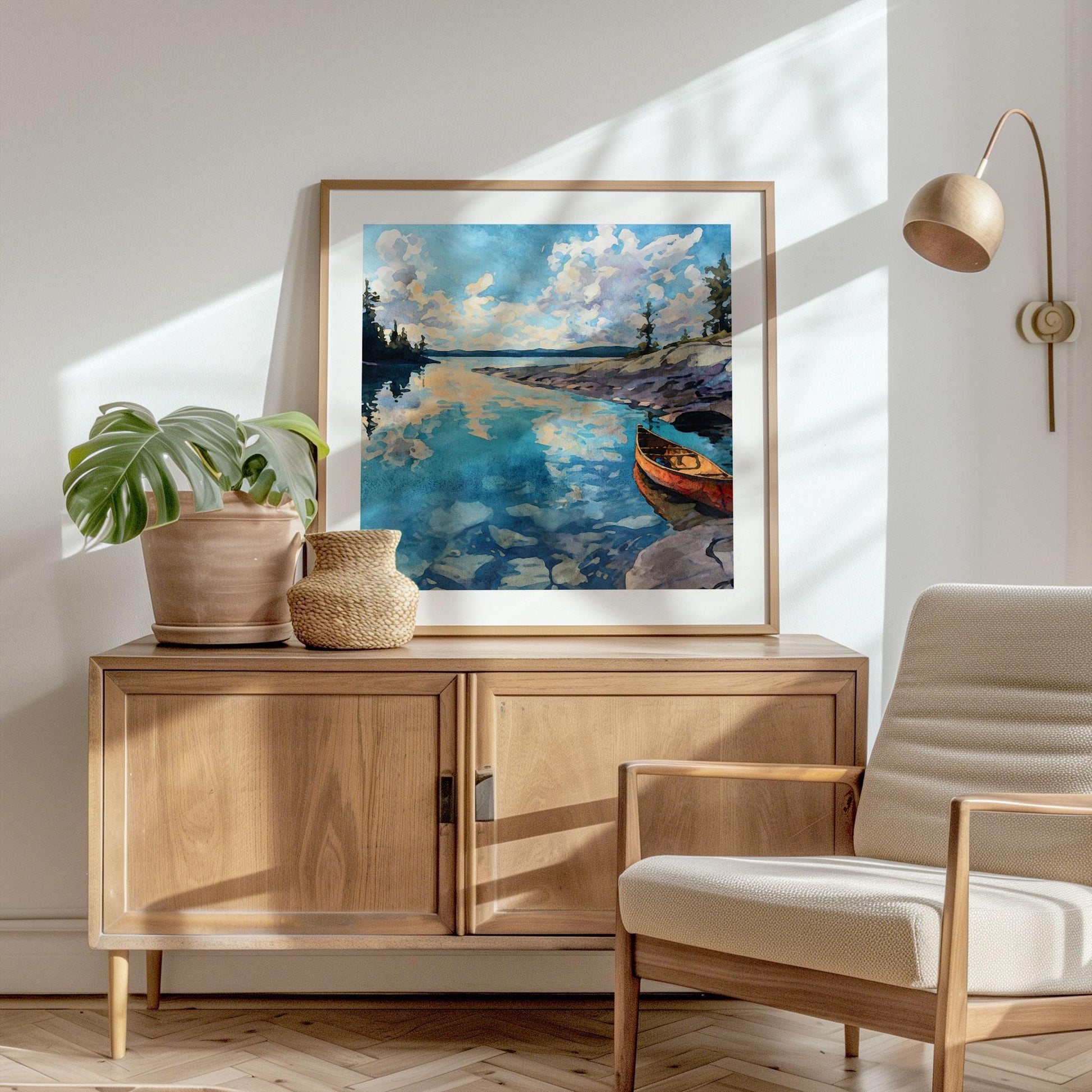 Framed square painting displayed on a wooden sideboard, depicting a red canoe resting by rocky shore with calm blue water reflecting clouds and pine trees. The scene is set in a bright, modern living room with a potted plant, woven vase, beige armchair, and warm sunlight streaming across the wall.
