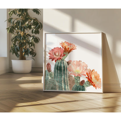 Watercolor cactus art print with pink, peach, and orange blooms in a white frame, leaning against a wall in a sunlit room with wood floors and a potted plant in the background.