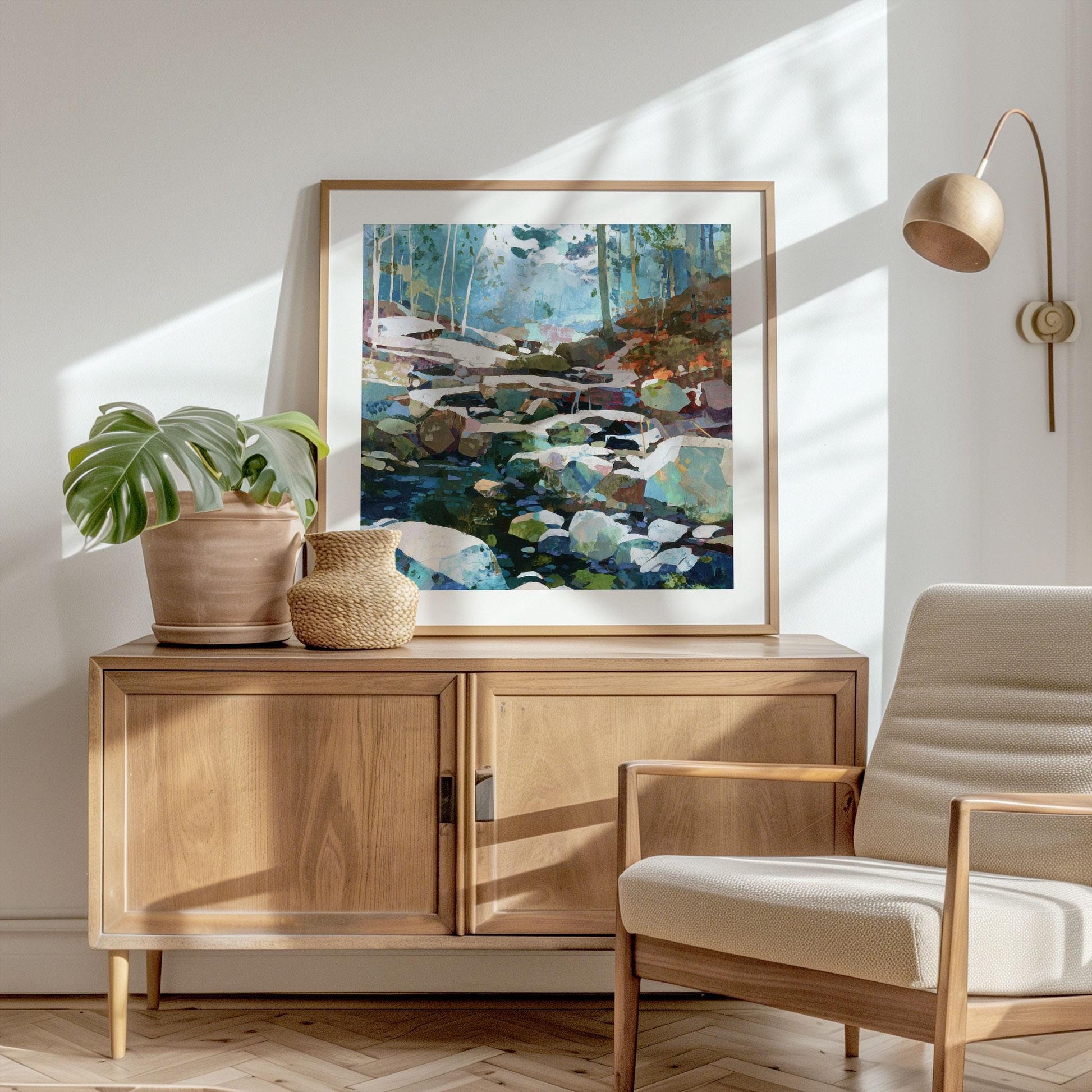 Framed square art print of a modern impressionist forest stream painting in cool blues, greens, and warm earth tones, displayed on a light wood sideboard. The minimalist room features a potted monstera plant, woven vase, beige armchair, and brass wall lamp, all bathed in soft natural sunlight.