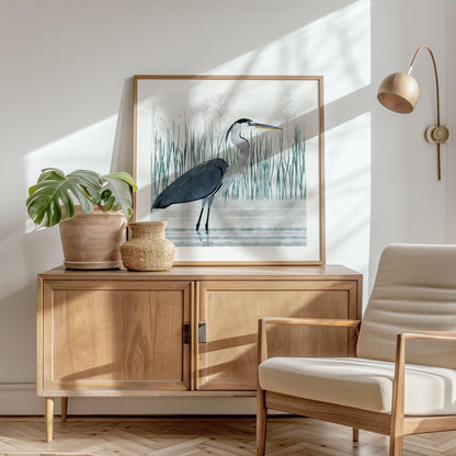 Framed art print of a great blue heron standing in water, resting on a light wood sideboard in a sunlit room. The heron is illustrated in a soft, watercolor style with tall reeds in the background. A potted monstera plant and woven vase sit beside the artwork, and a modern cream-colored armchair and wall-mounted lamp add to the room’s cozy, Scandinavian-inspired decor. Sunlight casts soft shadows across the white wall and parquet floor.