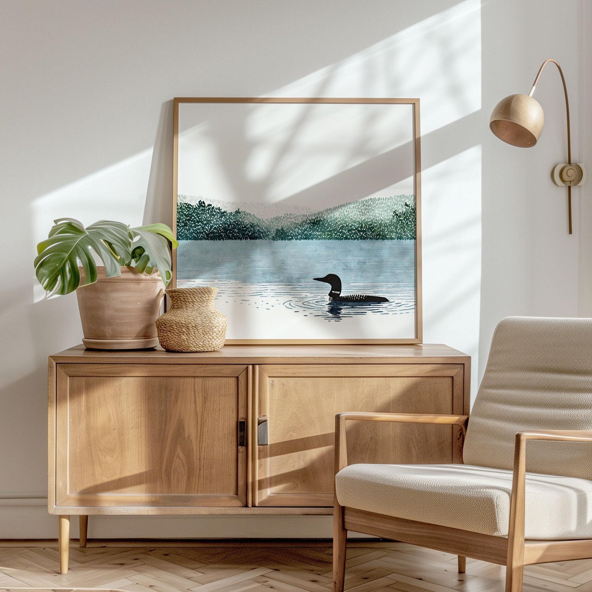 Framed loon art print leaning on a light wood cabinet, showing a black-and-white loon swimming on a tranquil blue lake with forested hills behind, styled in a sunlit room with a potted plant, woven basket, neutral armchair, and modern wall lamp.