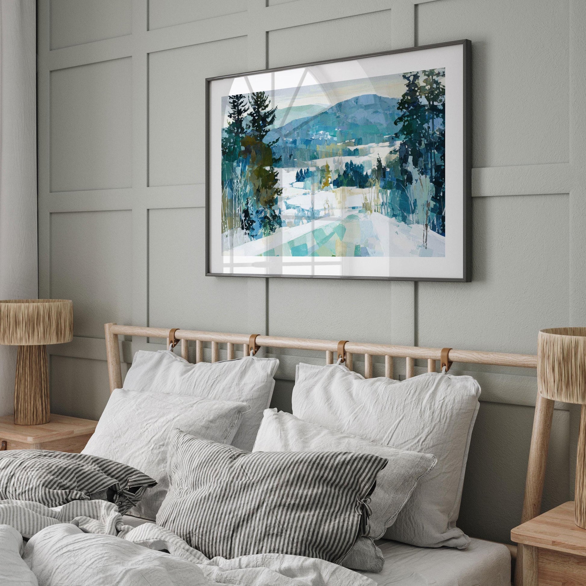 Framed winter landscape painting featuring snow, evergreen trees, and blue-green mountains, displayed above a bed with white linens and striped pillows in a modern, neutral-toned bedroom with wood accents and soft natural light.