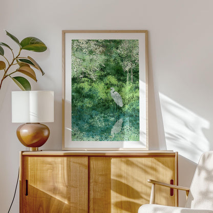 Framed watercolor-style art print featuring a white egret by a reflective pond, surrounded by lush green grasses and blossoming trees. Displayed in a sunlit modern room on a wooden cabinet beside a ceramic lamp and leafy plant, creating a calm, nature-inspired interior.