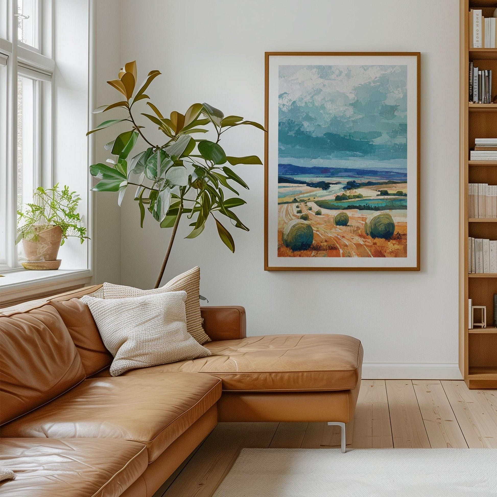 Modern living room with a tan leather sectional sofa, white throw pillows, and a potted plant by a window. On the wall hangs a large framed impressionist landscape painting of hay bales in golden fields under a dramatic teal sky.