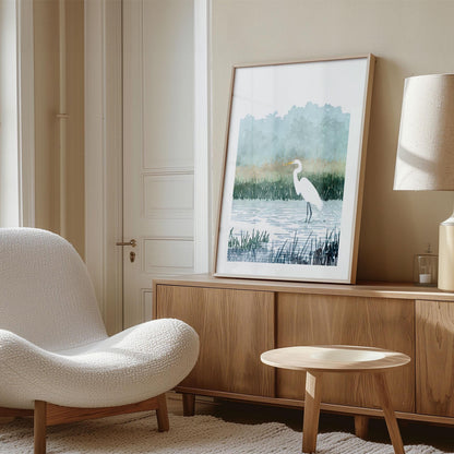 Framed watercolor-style print of a white egret standing in a marsh, leaning against the wall on top of a wooden sideboard in a modern living room. The room features a textured white lounge chair, round wooden coffee table, beige walls, and a neutral-toned lamp, creating a calm and minimalist aesthetic.