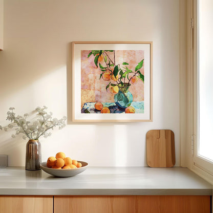 Square-framed still life painting of peaches in a blue glass vase, hung on a light kitchen wall near a window. The artwork shows leafy branches with ripe peaches against a textured pink background. Below, a countertop is styled with a ceramic vase of delicate white flowers, a wooden cutting board, and a bowl filled with fresh oranges, creating a bright and inviting scene.