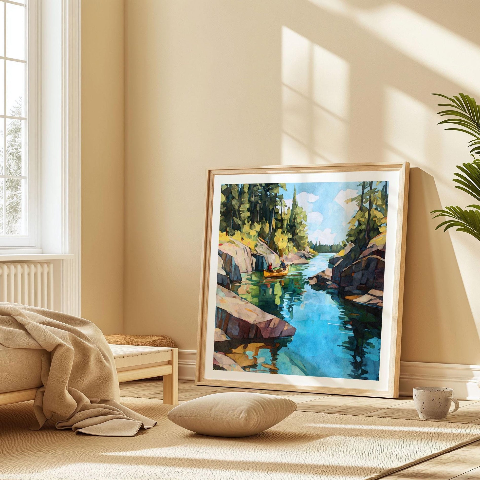 Large framed painting of a canoe on a calm lake between rocky cliffs and pine trees, leaning against a beige wall in a sunlit room with minimalist decor, soft bedding, a cushion, and a mug on the floor.
