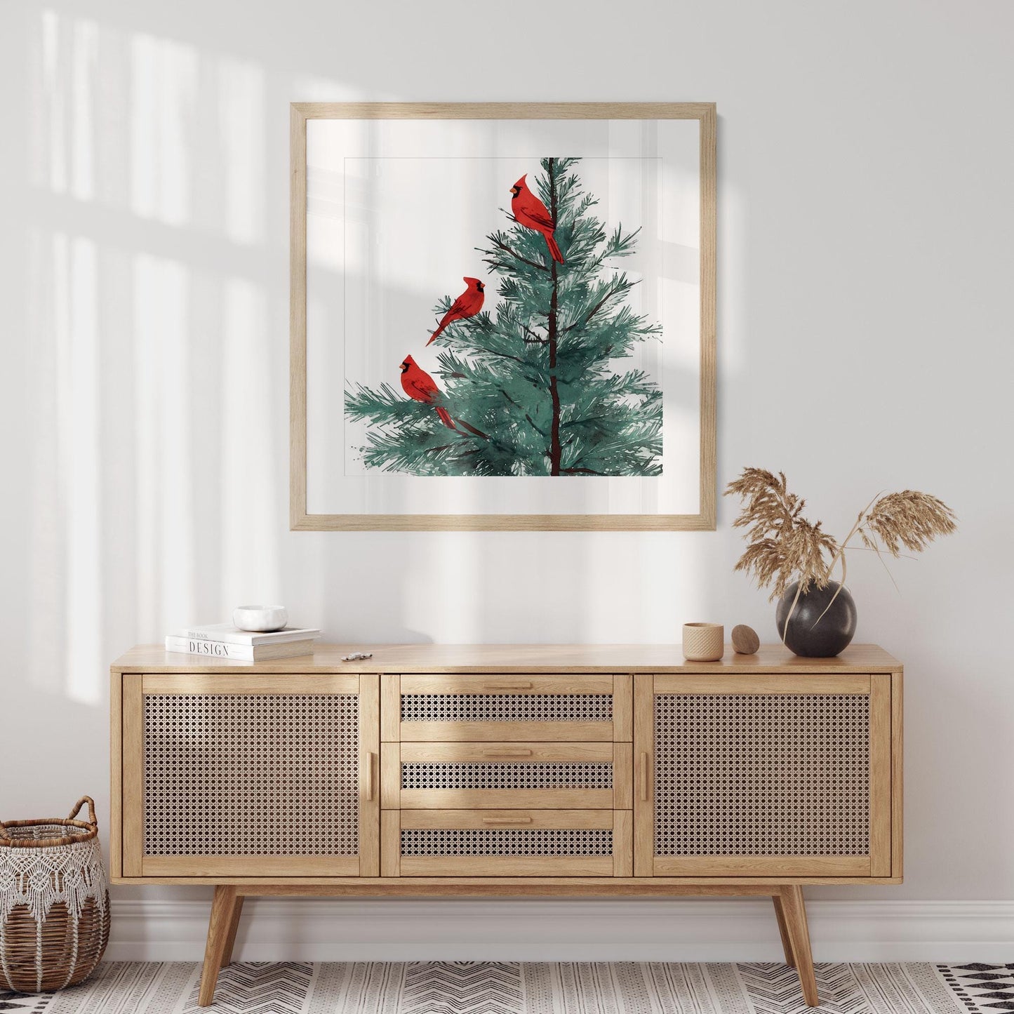 Framed art print of three red cardinals perched on a pine tree, hanging above a light wood sideboard with woven rattan doors. The modern room features neutral tones, a woven basket, books, and a black vase with dried foliage, creating a cozy minimalist aesthetic.