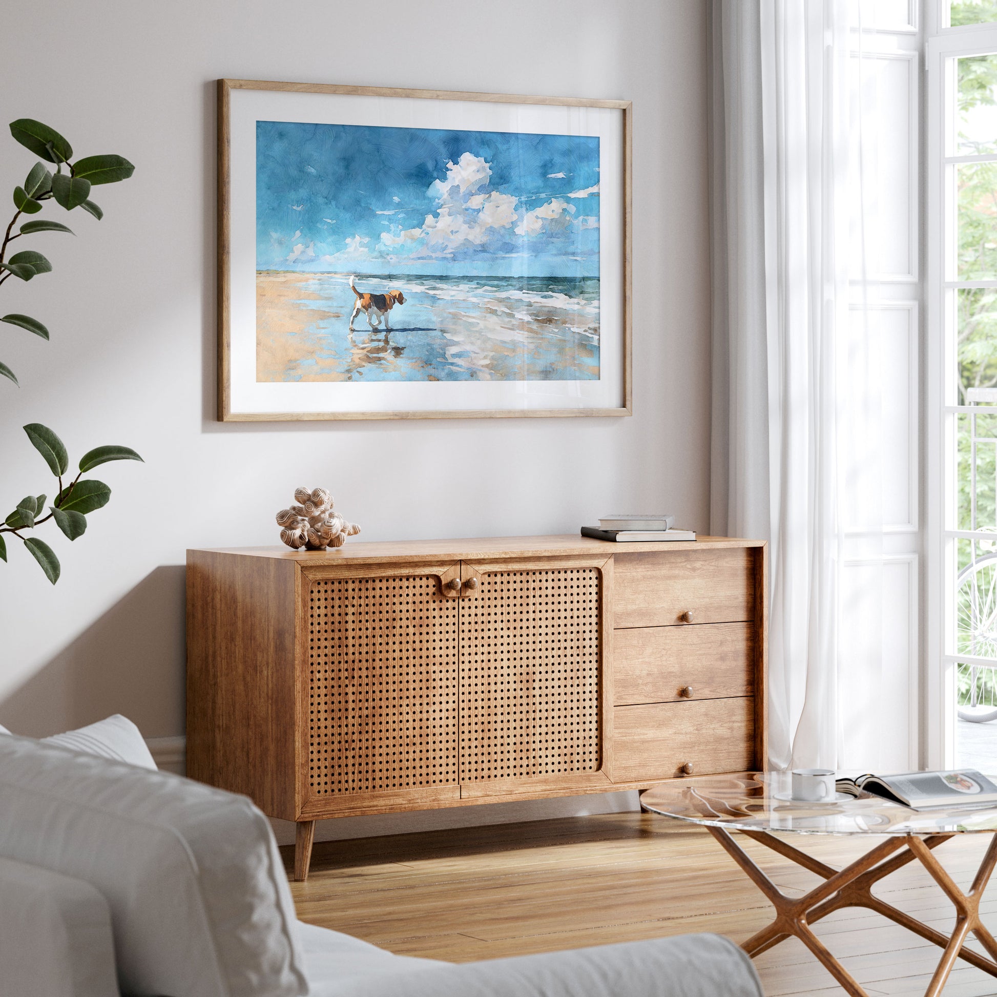 Framed watercolor-style beagle beach painting displayed above a mid-century wood sideboard in a bright, airy living room. The artwork shows a tricolor beagle walking along the shoreline with reflections in wet sand, beneath a blue sky with soft clouds, styled with neutral decor, plants, and natural light.