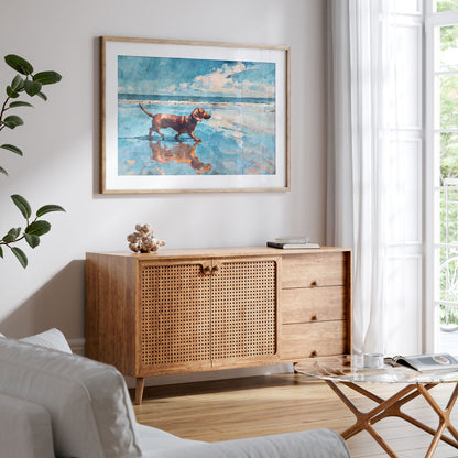 Framed watercolor-style beach scene of a red dachshund walking along the shoreline, reflected in wet sand under a bright blue sky. Displayed above a mid-century wood sideboard with cane doors in a light, airy living room with neutral tones and soft natural light.