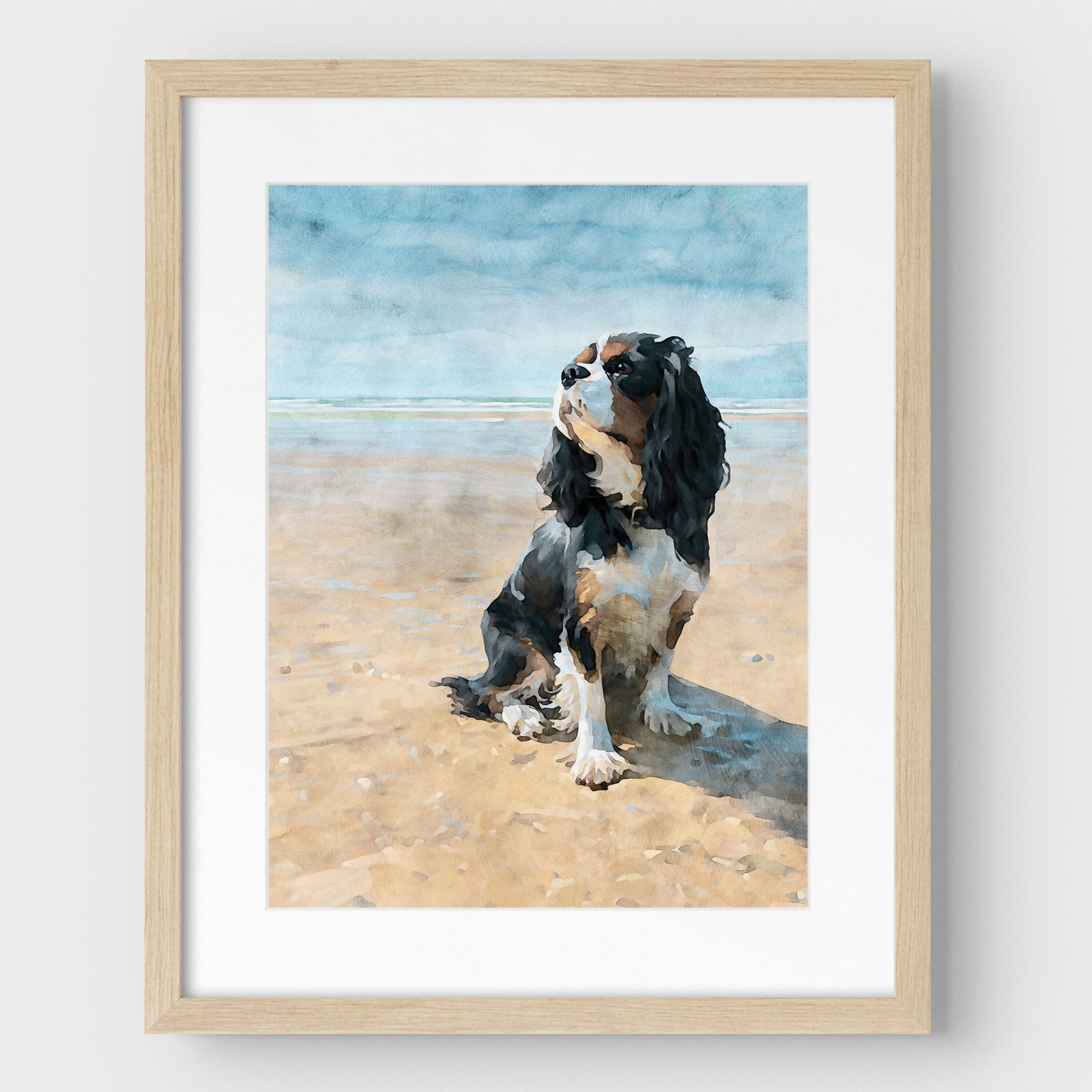 Framed artwork of a tricolor Cavalier King Charles spaniel on a beach with a light wood frame.