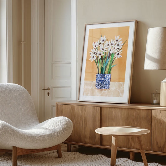 A framed print of white flowers with dark centers in a blue patterned vase, set against an orange background, displayed on a wooden sideboard in a bright room with neutral furnishings and soft natural light.