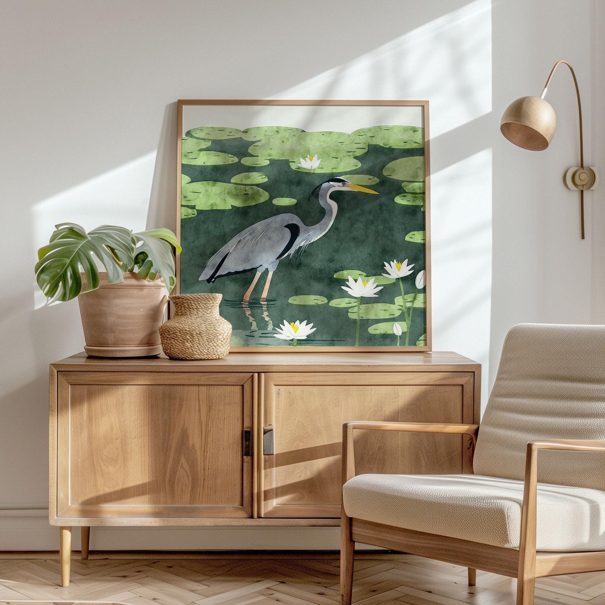 Framed heron art print featuring a blue-gray bird wading through a lily pad pond, with white lotus flowers and soft green leaves, in a watercolor-inspired style. Displayed atop a mid-century wooden cabinet in a sunlit room with a cream armchair, potted monstera, and rattan basket, the scene evokes a calm, natural elegance with warm shadows and minimalist decor.