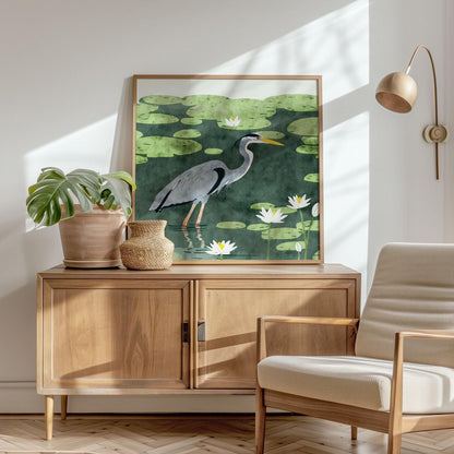 Framed heron art print featuring a blue-gray bird wading through a lily pad pond, with white lotus flowers and soft green leaves, in a watercolor-inspired style. Displayed atop a mid-century wooden cabinet in a sunlit room with a cream armchair, potted monstera, and rattan basket, the scene evokes a calm, natural elegance with warm shadows and minimalist decor.