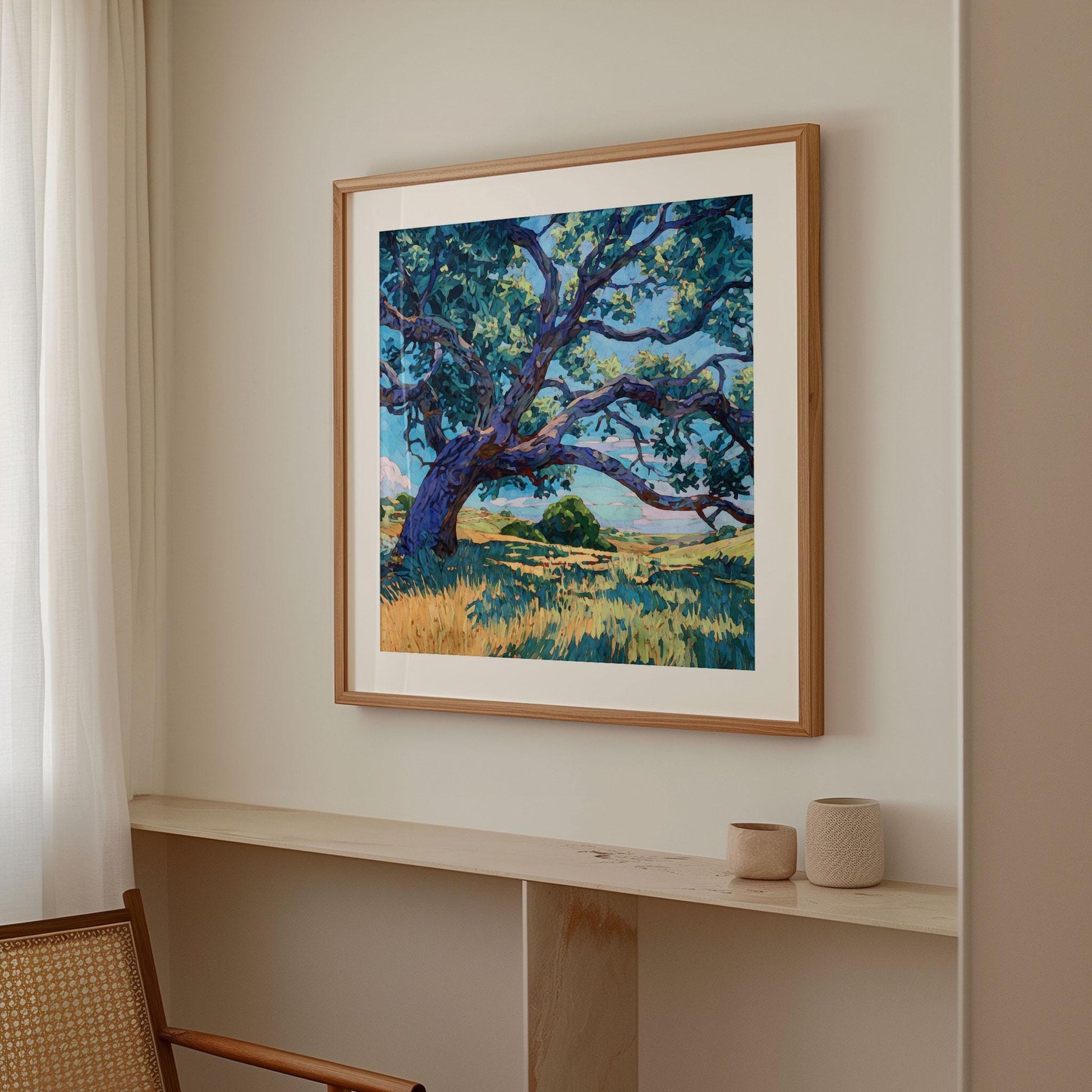Framed art print of a sprawling Texas oak tree hangs on a neutral beige wall beside a softly draped white curtain. The modern impressionist painting features twisting blue-violet branches over golden grass under a bright blue sky. A cane chair and two small textured ceramic vessels rest on a light wood ledge, creating a calm, minimalist space with warm natural light.