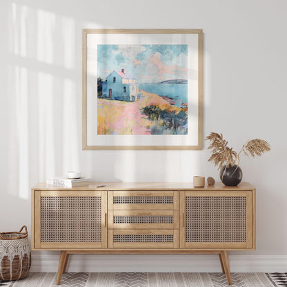 Square framed watercolor painting of a coastal scene with a light blue house on a bluff overlooking the ocean, hung above a light wood cabinet with woven rattan doors. The room is styled with a black vase holding dried grasses, a small basket on the floor, and books on the cabinet, creating a warm, airy, and modern coastal aesthetic.