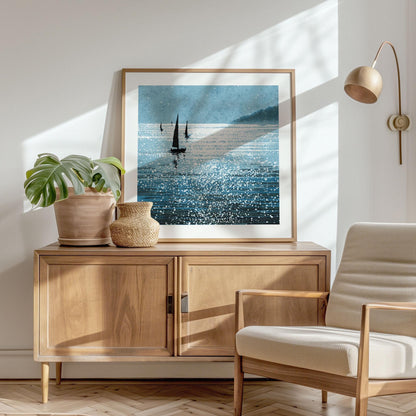 Square-format sailboat art print in a light wood frame, leaning against a wall atop a mid-century modern cabinet. The artwork features dark sailboats on sparkling blue water, with sunlight dancing across the surface and a hazy headland in the distance. Framed with a white matte border, it’s styled with a potted monstera, woven vase, and cream armchair in a sunlit, minimalist interior.