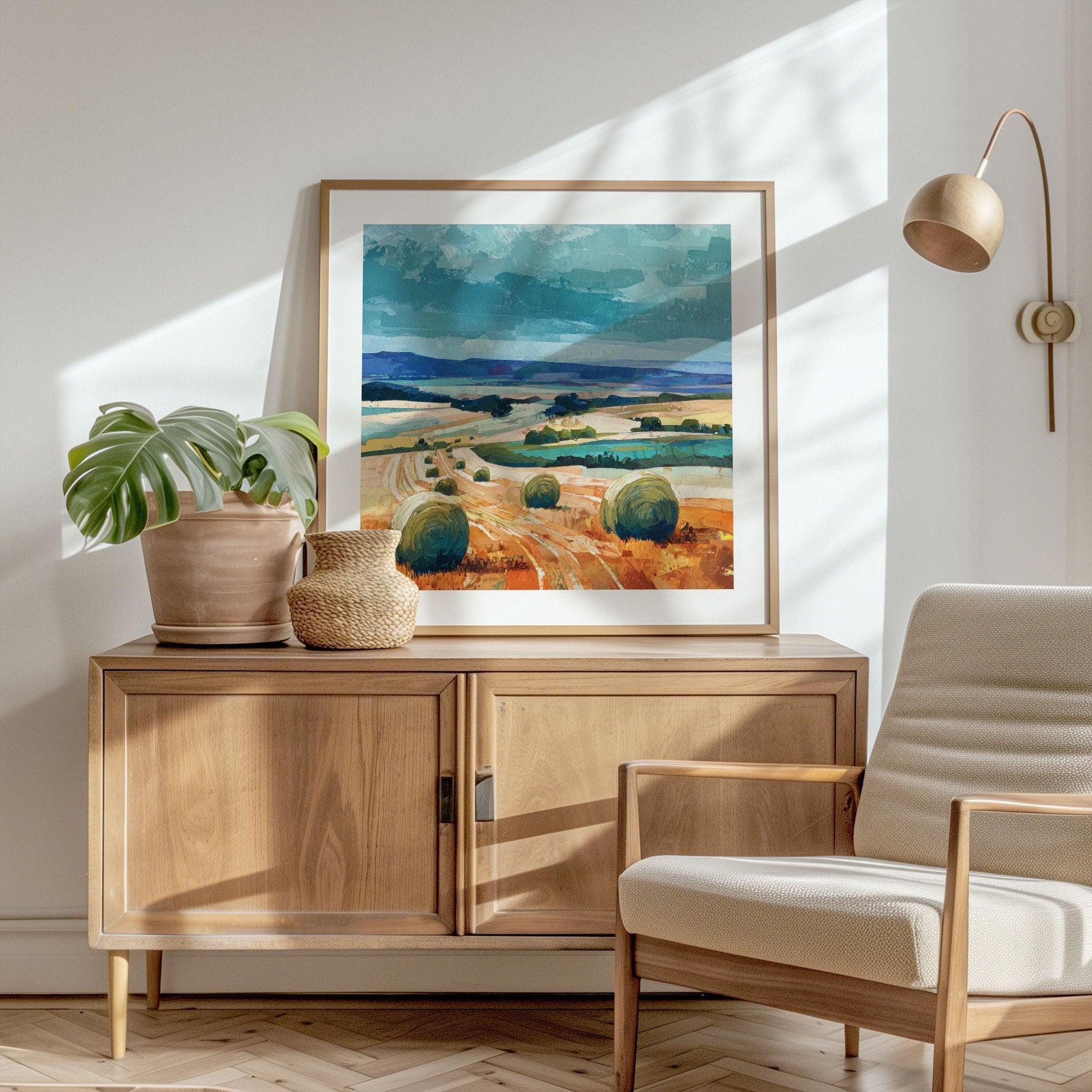 Framed impressionist countryside painting of hay bales in golden fields under a turquoise sky, displayed on a light wood cabinet. The scene is styled in a modern room with a potted monstera, woven basket, cream armchair, and brass wall lamp.