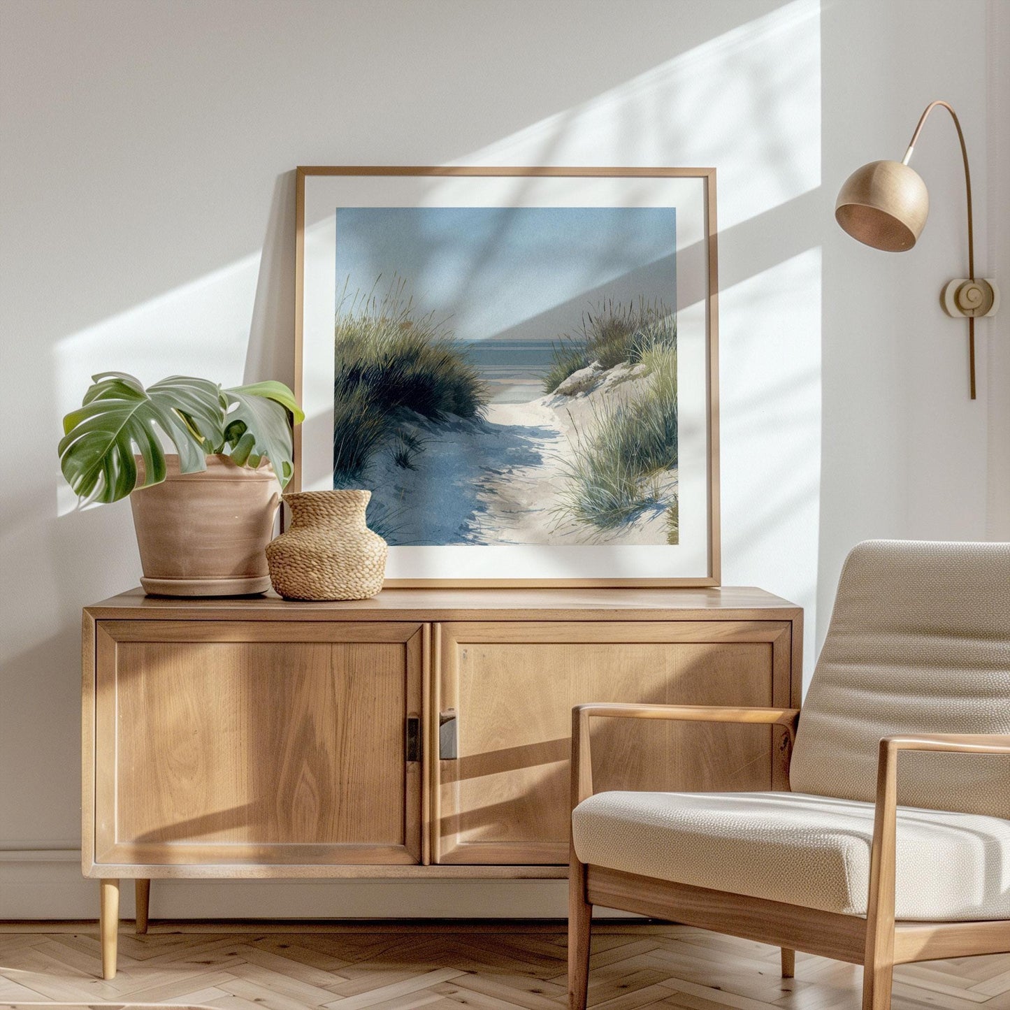 Framed coastal landscape print of a sandy beach path through tall grass, styled in soft watercolor tones, displayed on a wooden sideboard. Decor includes a potted monstera plant, woven basket, neutral armchair, and wall-mounted lamp in a bright, sunlit room.