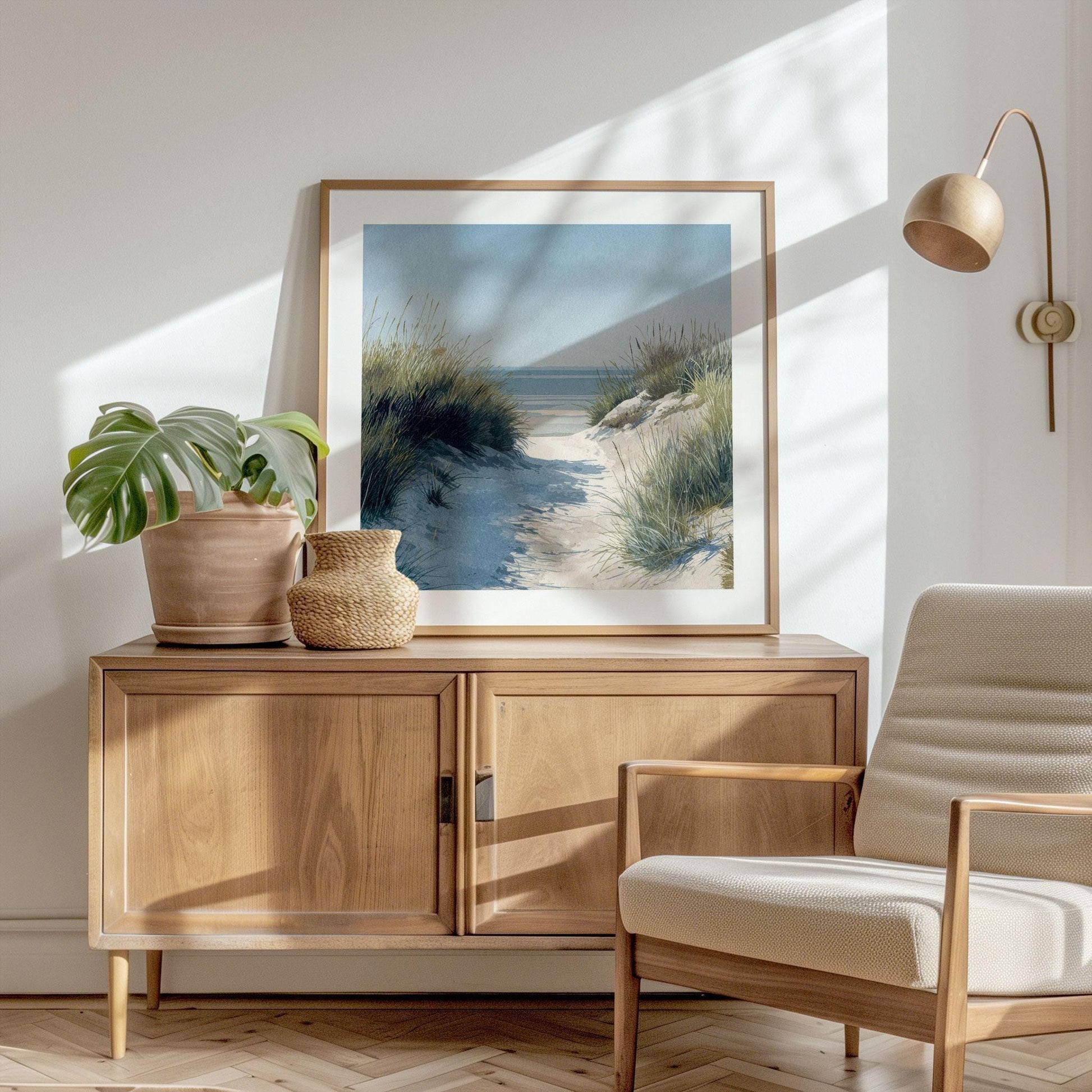 Framed coastal landscape print of a sandy beach path through tall grass, styled in soft watercolor tones, displayed on a wooden sideboard. Decor includes a potted monstera plant, woven basket, neutral armchair, and wall-mounted lamp in a bright, sunlit room.