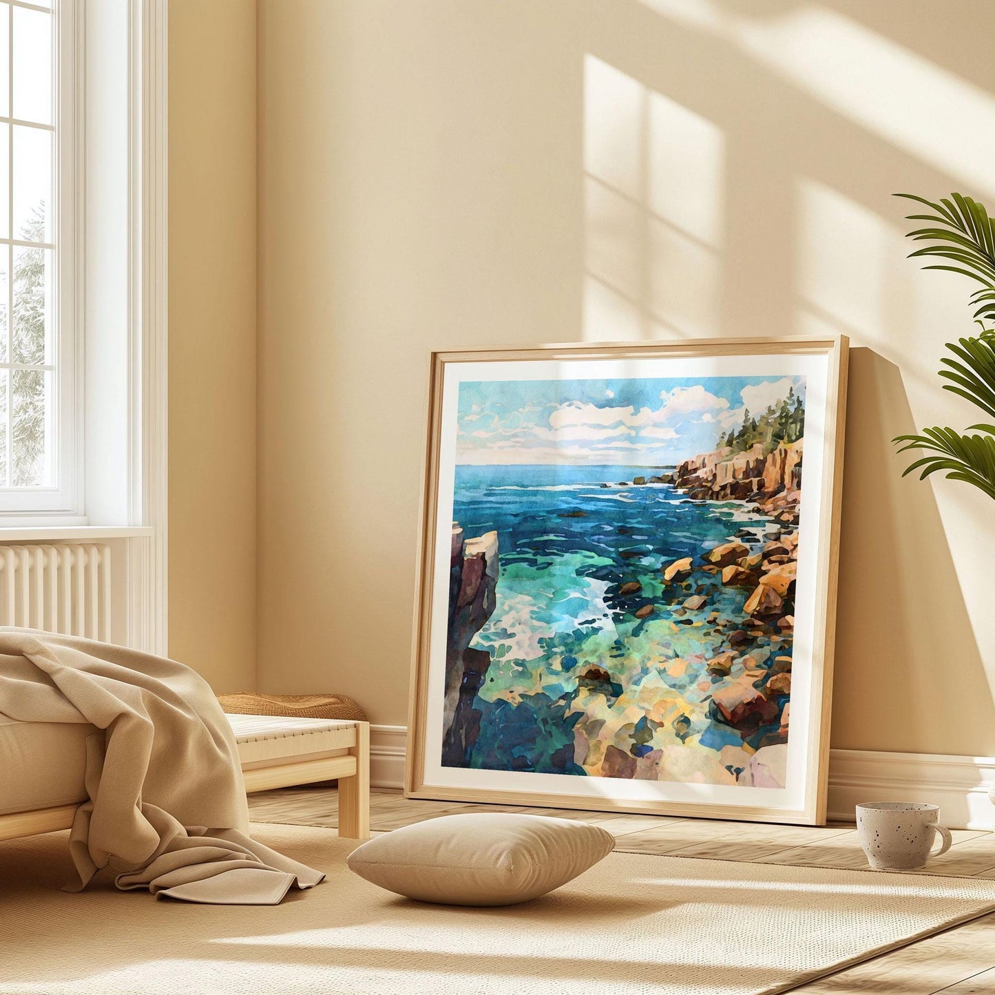 Framed watercolor-style coastal artwork leaning against a beige wall in a sunlit minimalist room. The painting depicts rocky cliffs, turquoise waters, and a bright sky with clouds. The frame rests on a light rug beside a low bed with a tan blanket, a floor cushion, a mug, and a potted plant, creating a serene, cozy atmosphere.