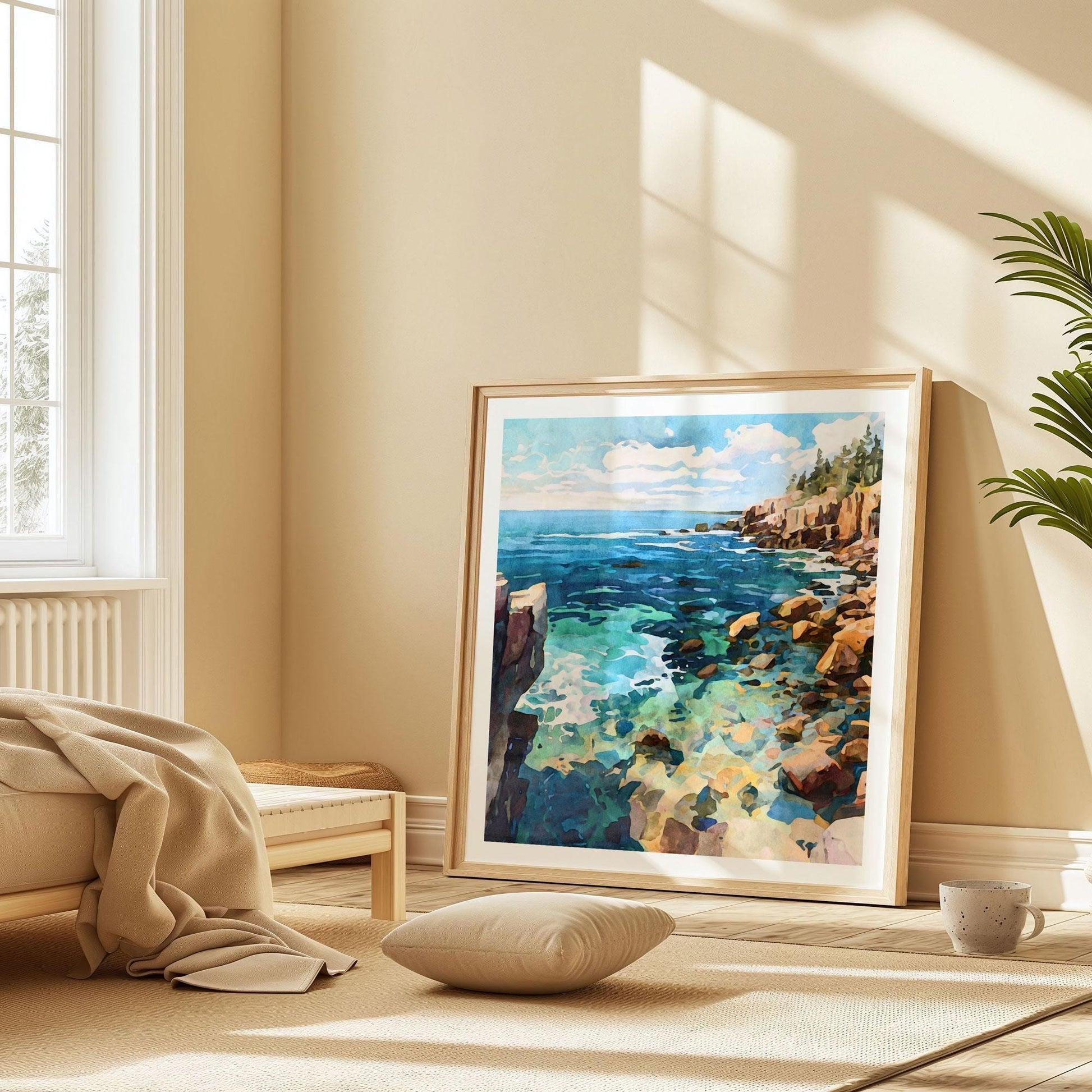 Framed watercolor-style coastal artwork leaning against a beige wall in a sunlit minimalist room. The painting depicts rocky cliffs, turquoise waters, and a bright sky with clouds. The frame rests on a light rug beside a low bed with a tan blanket, a floor cushion, a mug, and a potted plant, creating a serene, cozy atmosphere.