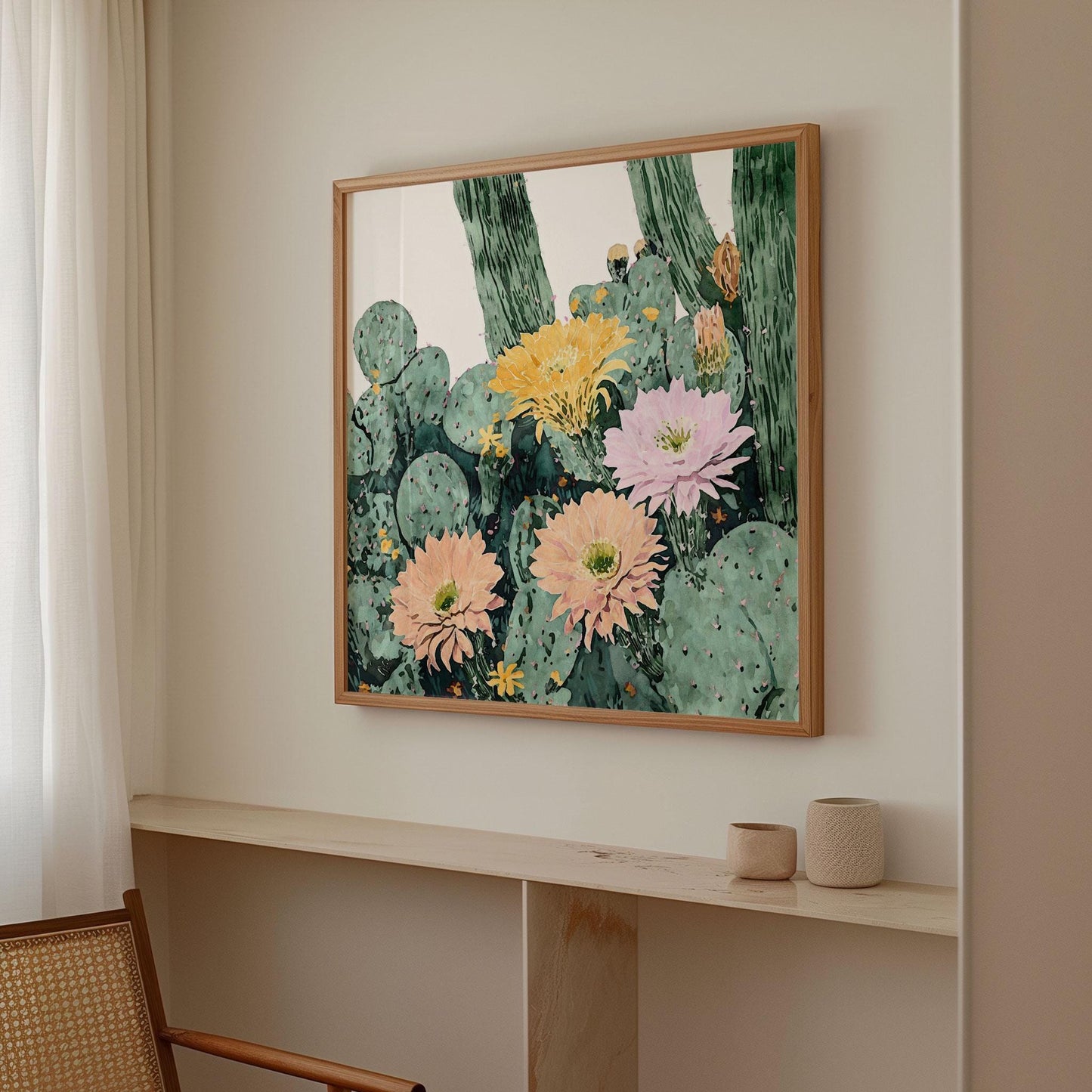 Framed botanical art print featuring watercolor-style cacti with large peach, pink, and yellow blooms, hanging on a neutral-toned wall in a minimalist room. The space includes sheer curtains, a woven chair, and small ceramic vessels on a light wood shelf, creating a calm and airy atmosphere.