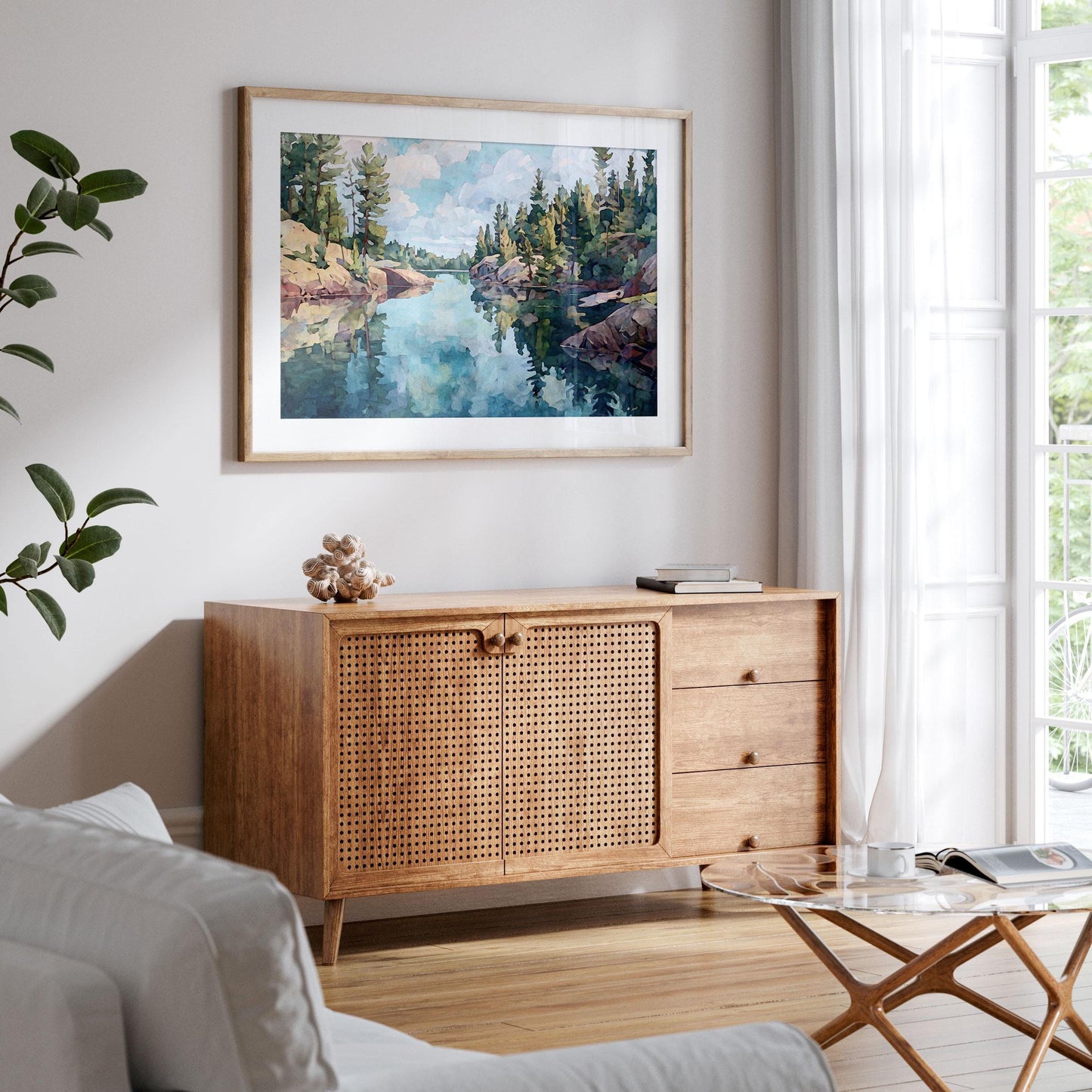Framed landscape painting of a northern Minnesota lake hangs above a mid-century style wood sideboard with woven rattan doors. The artwork shows calm blue-green water reflecting pine trees and rocky shores under a cloudy sky. Room features white walls, a light sofa, glass coffee table, and a large window with sheer curtains, creating a bright, airy, modern space.